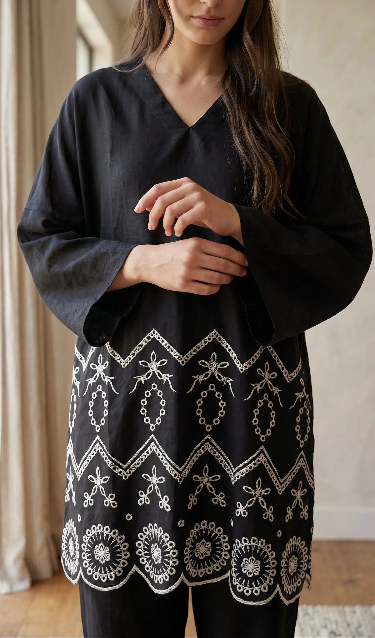 A woman wearing a loose black tunic top with white embroidered geometric and floral patterns along the hem, standing indoors against a neutral beige wall.
