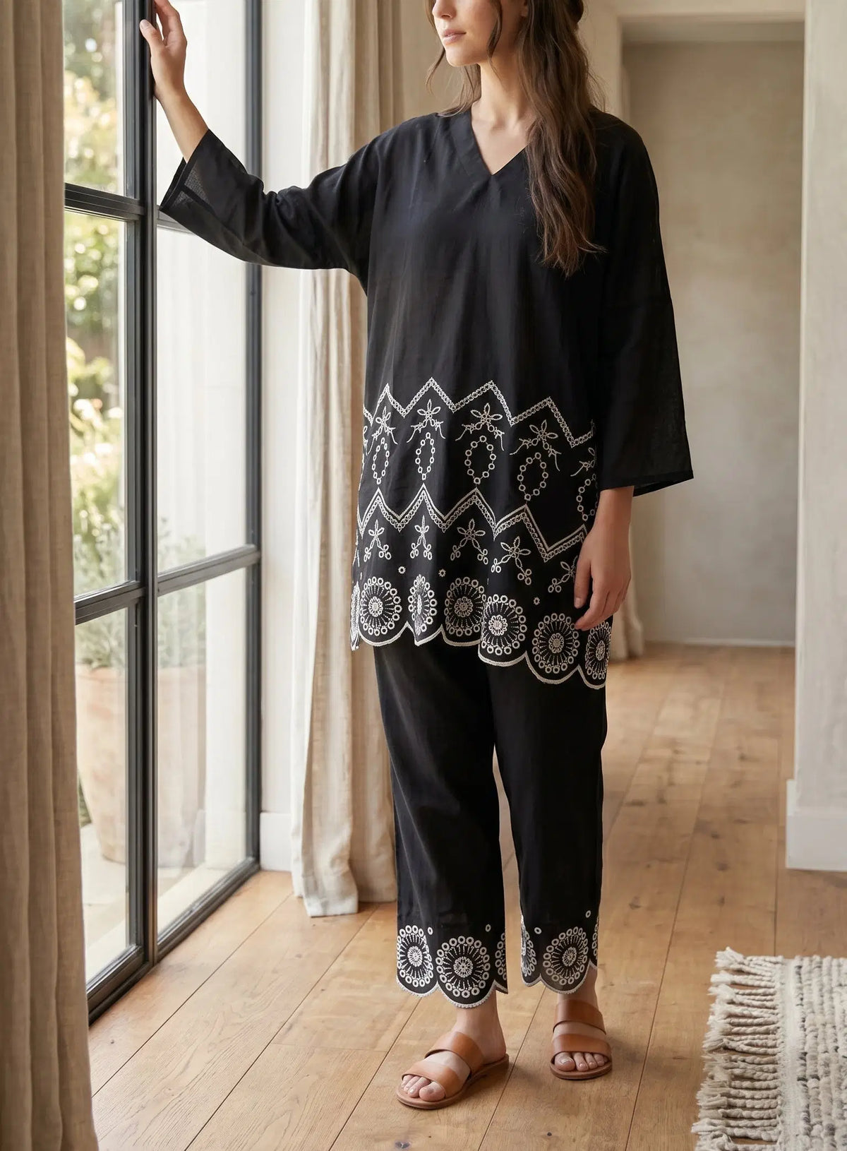 A woman wearing a black two-piece tunic and trouser set with intricate white embroidered patterns stands by a large window in a bright room with wooden floors.