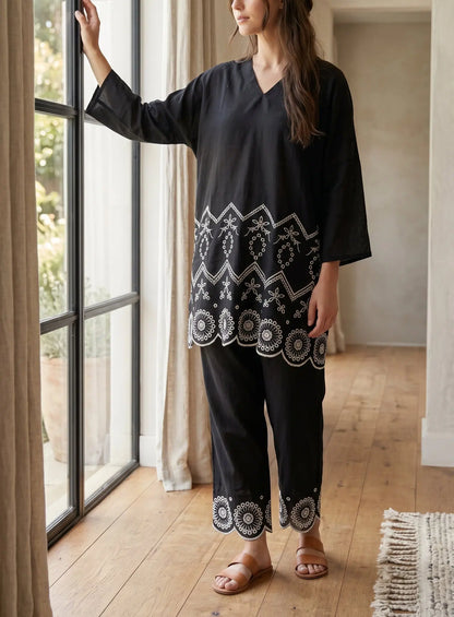 A woman wearing a black two-piece tunic and trouser set with intricate white embroidered patterns stands by a large window in a bright room with wooden floors.