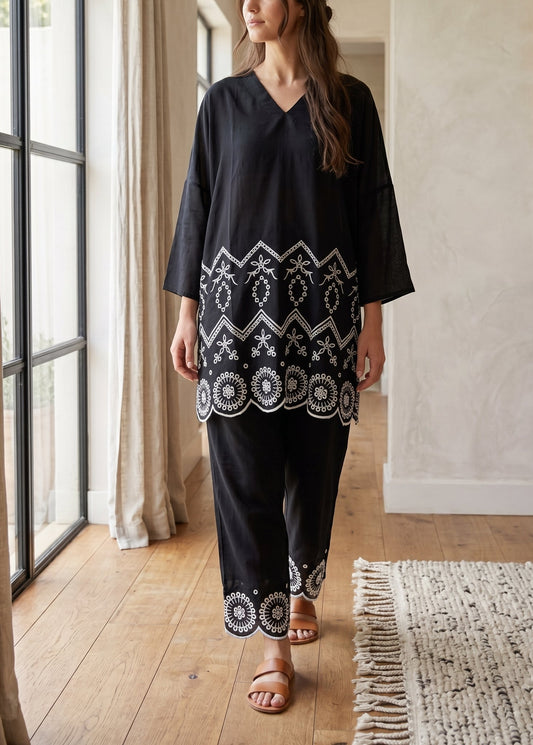 A woman modeling a black two-piece tunic and trouser set with white geometric embroidery, standing in a bright room with wooden floors and large windows.