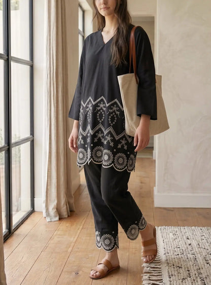 A woman modeling a black tunic and pants set with intricate white embroidery, standing in a bright room with wooden floors and large windows.